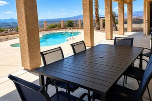 Un espace piscine extérieur avec une table et des chaises, entouré d’une clôture et de palmiers, offrant une vue sur les montagnes en arrière-plan.