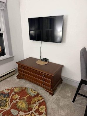 A room with a flat-screen TV mounted on the wall, a wooden dresser with drawers, a patterned rug, and a chair. - NEW Listing Half Block to Downtown Treetop Apt 3bd (Black Mountain)