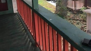 En overdækket veranda med rødt gelænder, et træ i baggrunden og en indhegnet have.