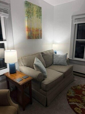 A living room with a sofa, a side table with a lamp, and a painting on the wall. - NEW Listing Half Block to Downtown Treetop Apt 3bd (Black Mountain)