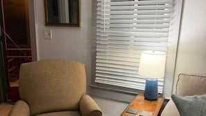 En stue med en beige lænestol, et lille træbord med en lampe og et vindue med persienner.