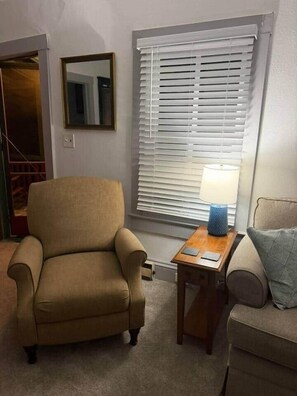 A living room with a beige armchair, a small wooden table with a lamp, and a window with blinds. - NEW Listing Half Block to Downtown Treetop Apt 3bd (Black Mountain)