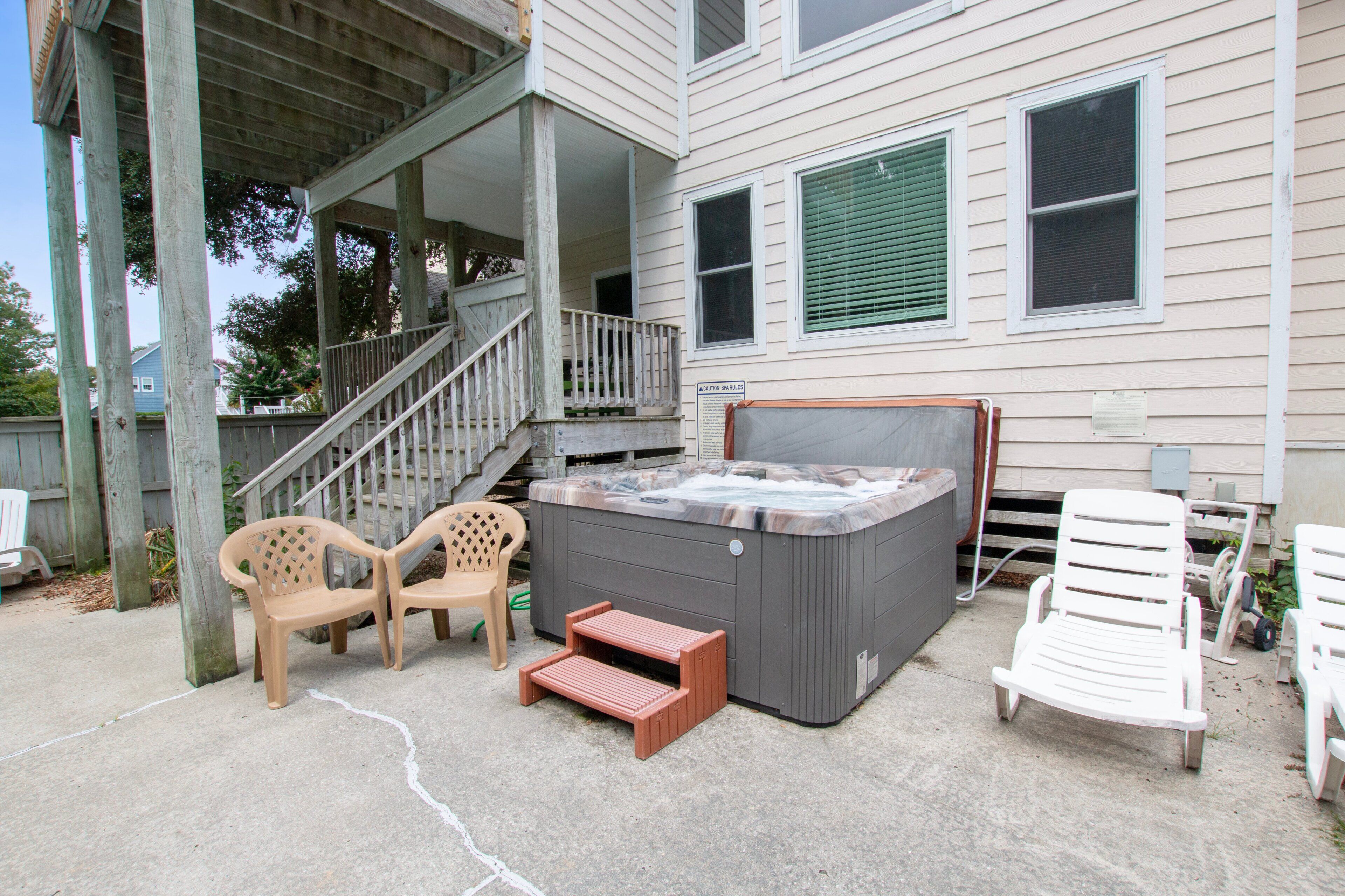 A hot tub with a wooden deck, outdoor chairs, and a staircase leading to a porch.