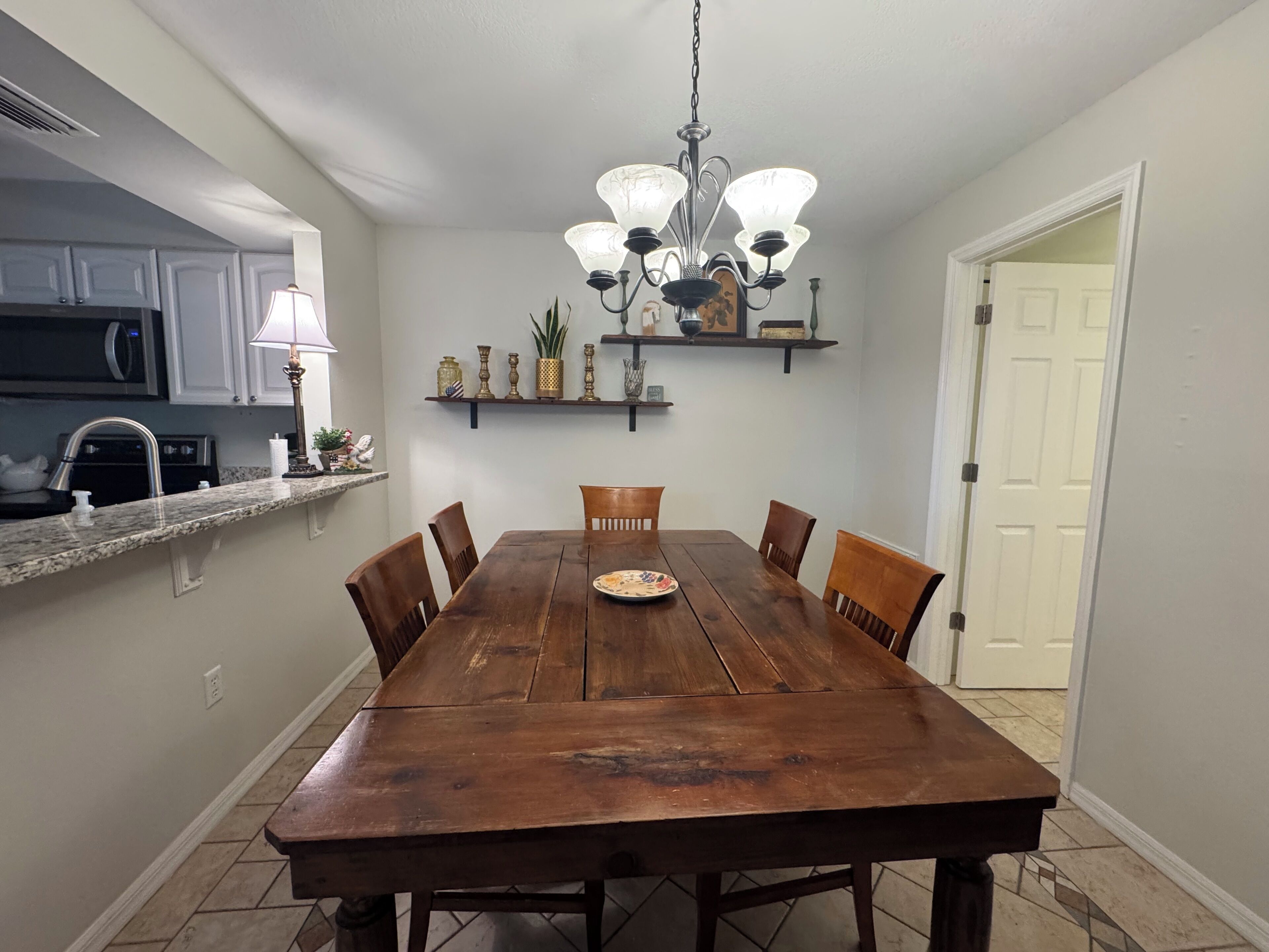 A dining area with a wooden table, chairs, a chandelier, and a kitchen in the background.