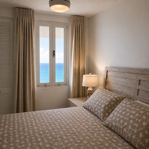 A hotel room with a bed, a nightstand with a lamp, and a window with curtains. - Oceanfront 1BR Condo with Stunning Beach Views & AC (Luquillo)