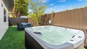 Un patio trasero con jacuzzi, césped artificial, valla de madera y una mesa con sillas.