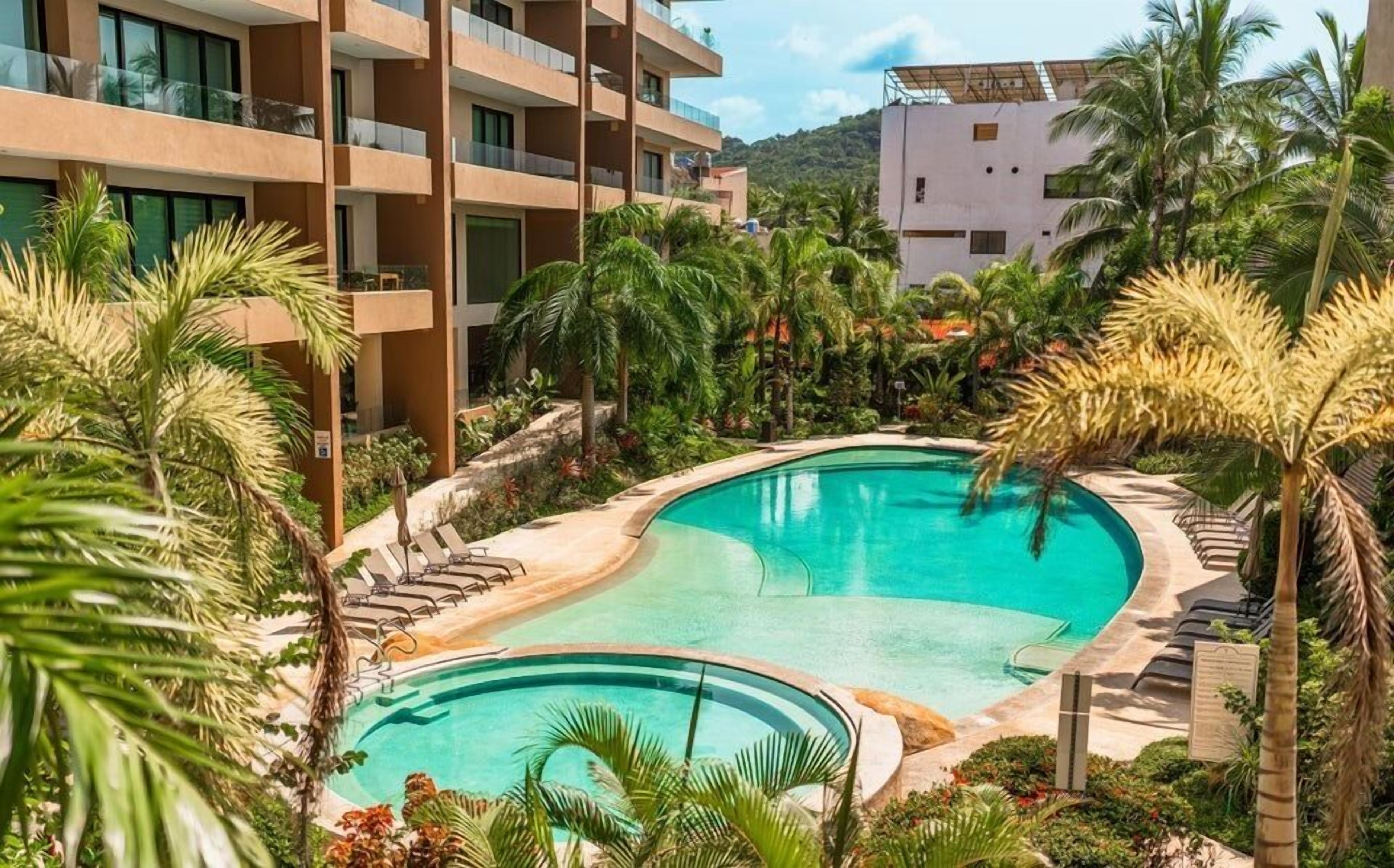 Un resort con una amplia piscina rodeada de palmeras y un edificio moderno con balcones.