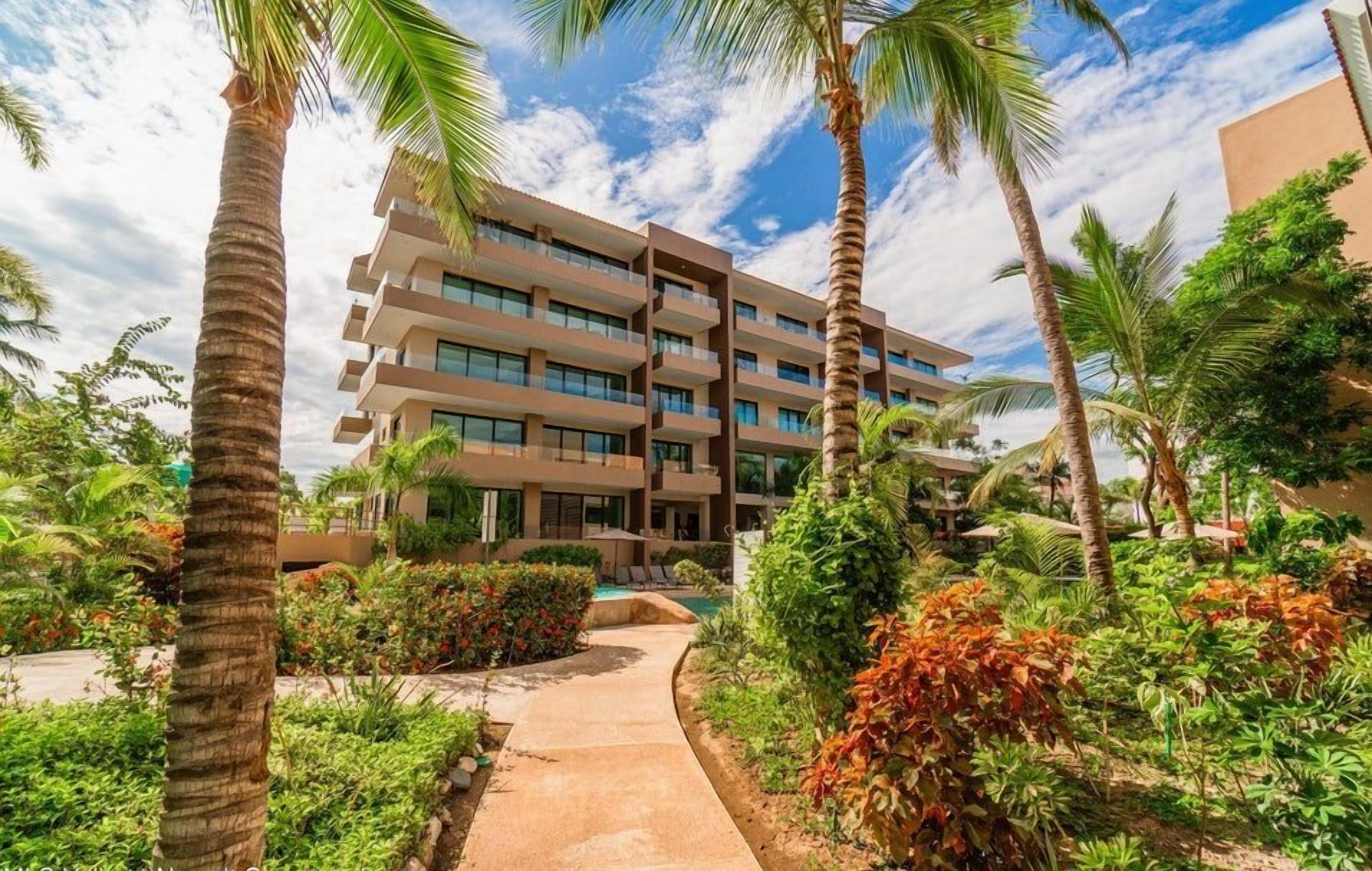Un hotel de varios pisos con balcones, rodeado de abundante vegetación y palmeras.