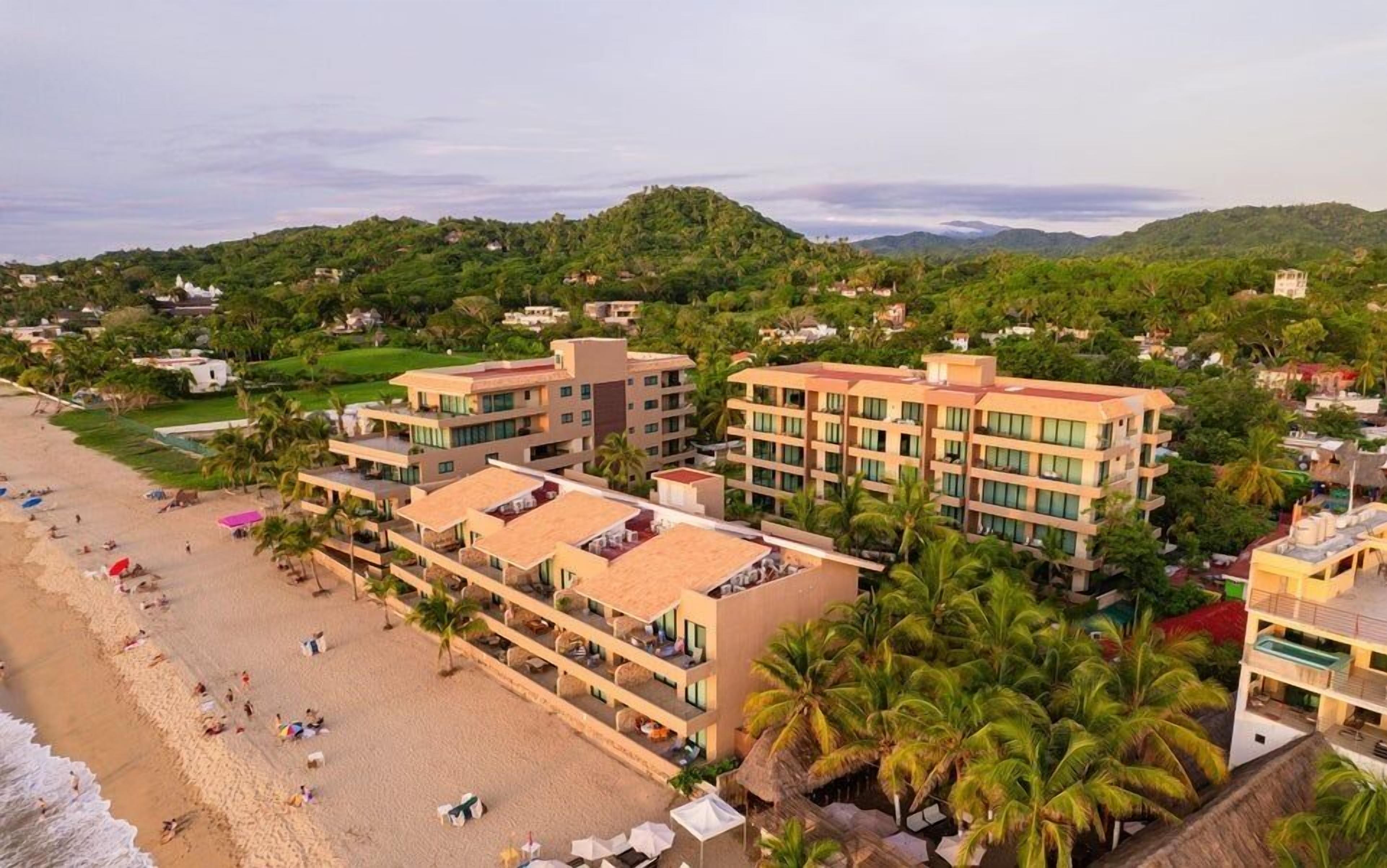 Un hotel frente a la playa con varias construcciones, una playa de arena y palmeras.