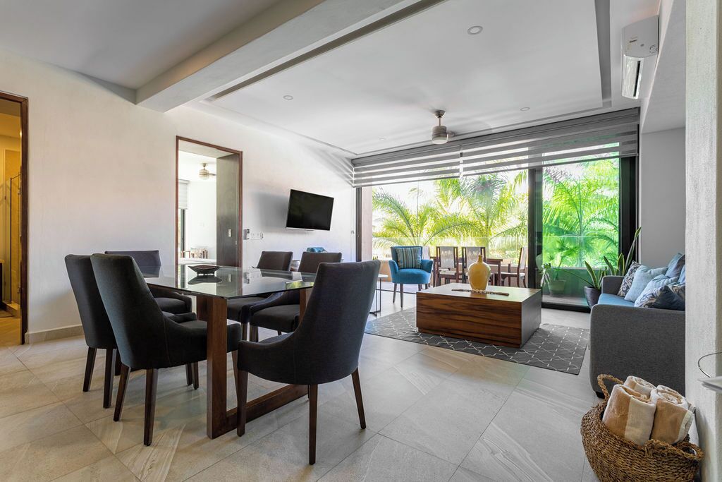 A modern living room with a dining area, large windows, and a view of greenery.