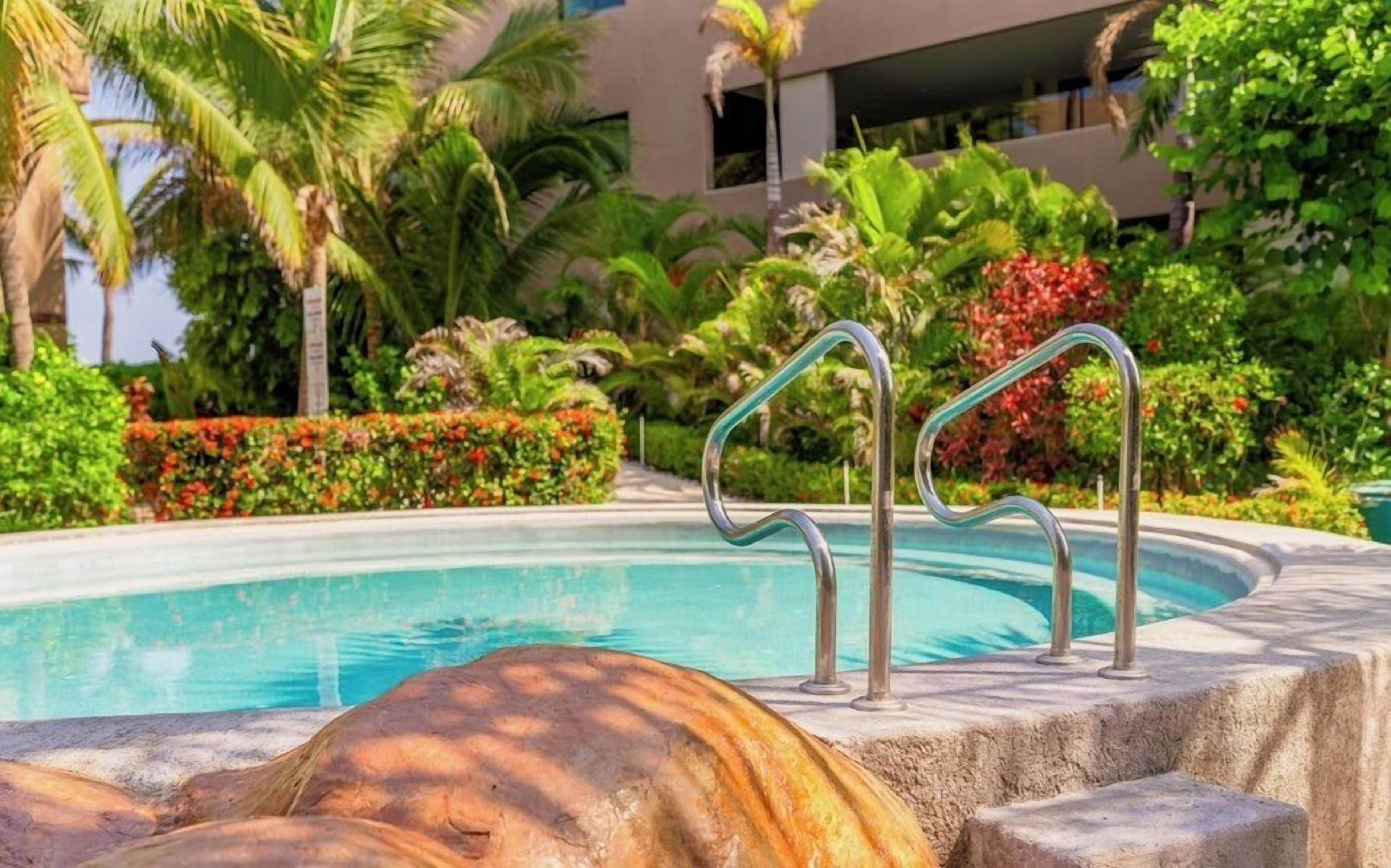 A swimming pool with metal handrails, surrounded by lush greenery and palm trees.