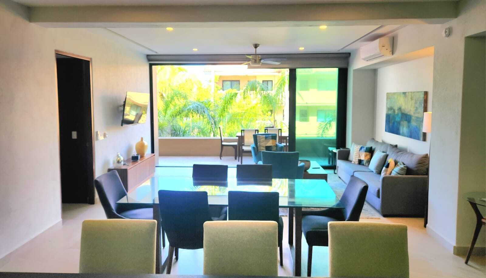 A modern living room with a glass dining area, a sofa set, and a flat-screen TV mounted on the wall.