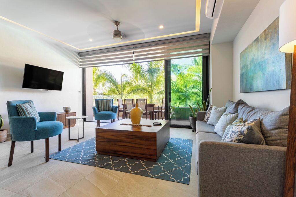 A modern living room with a flat-screen TV, a wooden coffee table, a blue armchair, a grey sofa, and a large window with a view of palm trees.