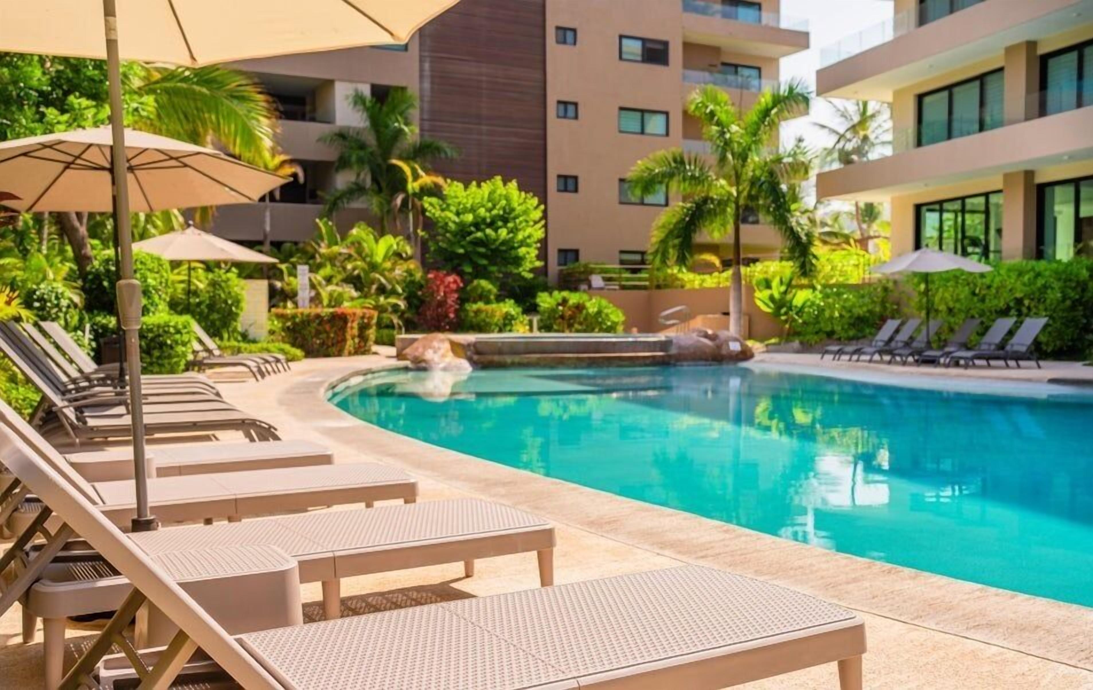 A pool area with lounge chairs and umbrellas, surrounded by greenery and a modern building.