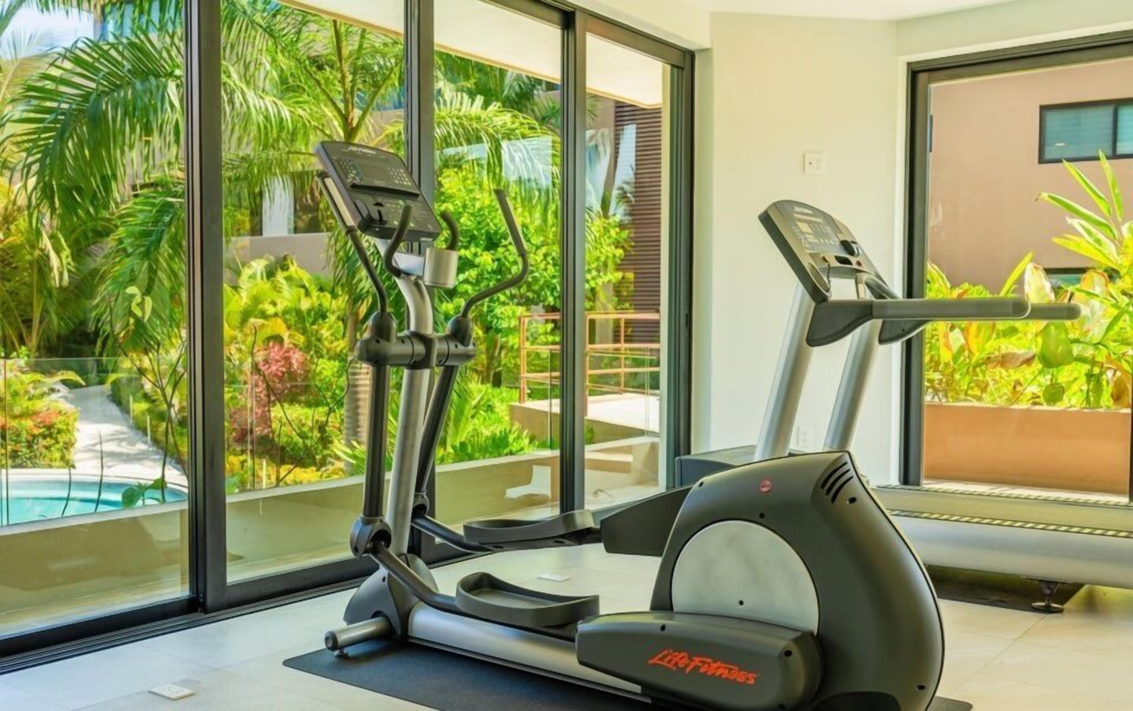 Gimnasio en casa con equipo cardiovascular, una cinta de correr y una máquina elíptica, con vistas a una piscina y exuberante vegetación.