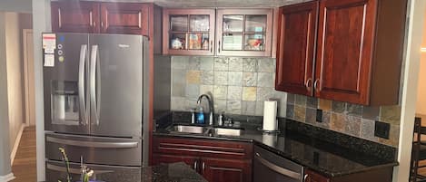 A modern kitchen with dark wood cabinets, a stainless steel refrigerator, a granite countertop, and a built-in dishwasher.