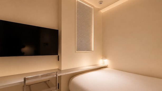 Quarto com cama, televisão presa na parede, janela com persianas e mesa de cabeceira com abajur.