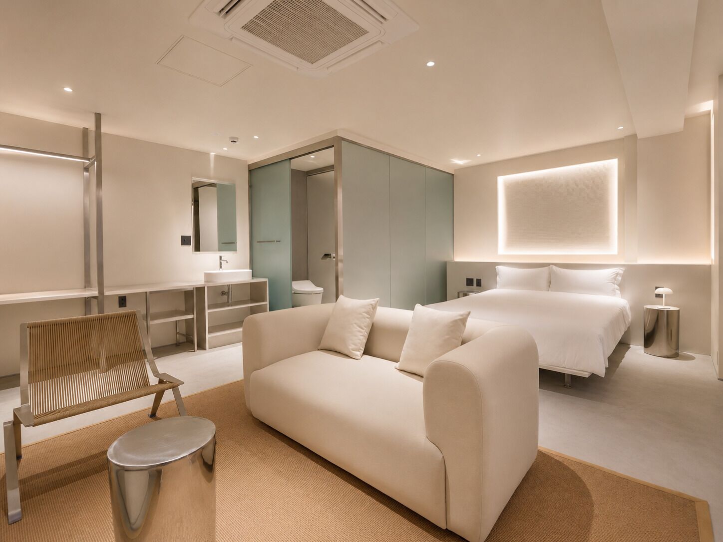 A modern hotel room with a sofa, a small round table, a bed, a nightstand, and a bathroom with a mirror and sink.