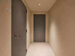 A hallway with a closed door at the end, a door with a handle on the left, and a light fixture on the ceiling. - TORA HOTEL Ueno 5-chome  (Tokyo)