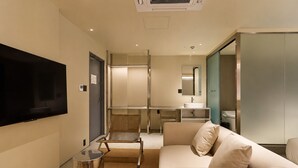 Quarto de hotel moderno com televisão de tela plana, sofá, uma mesinha e banheiro com pia e espelho.