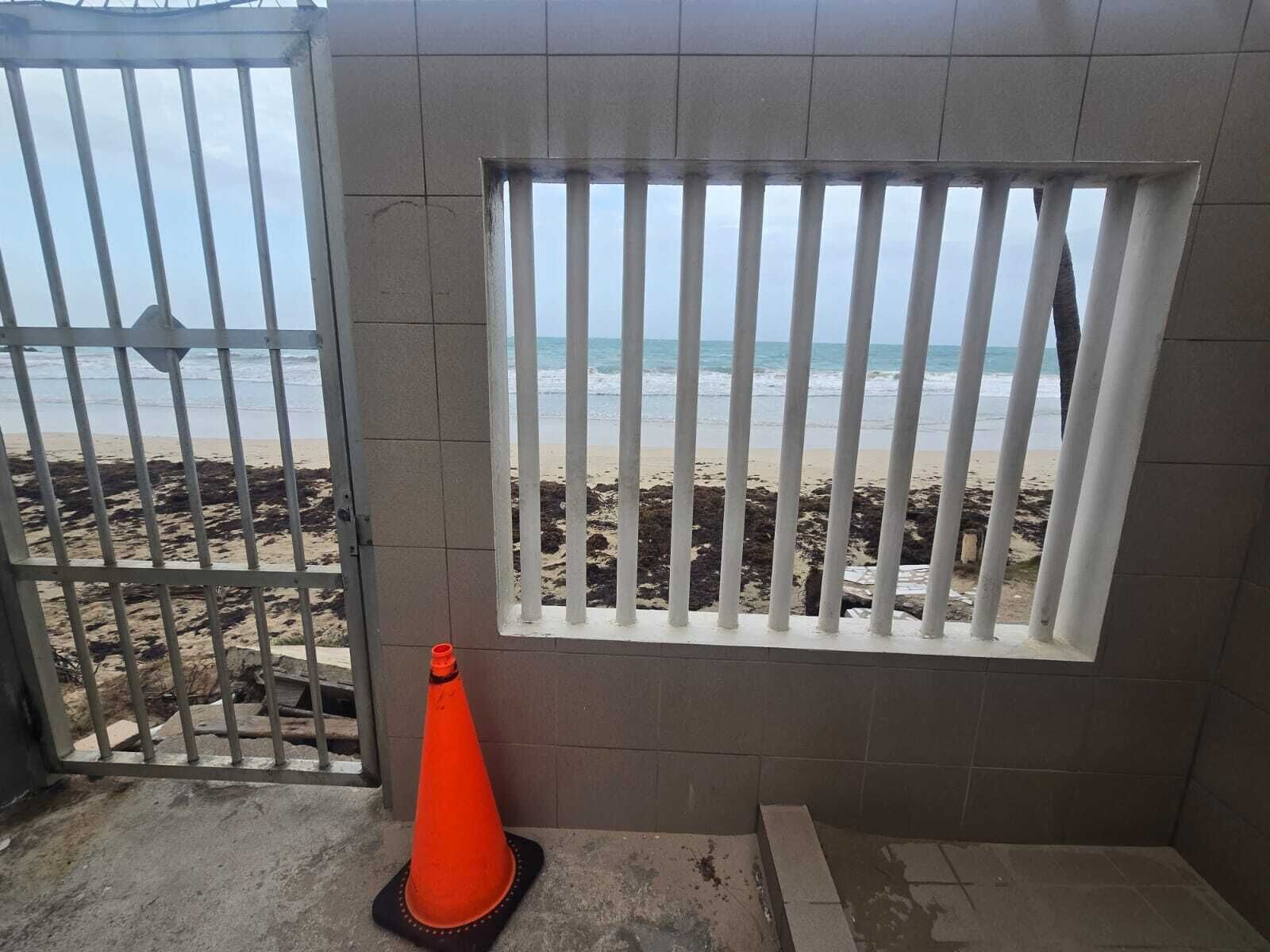 Blick auf einen Strand durch ein vergittertes Fenster, ein Betonboden und ein einzelner, orangefarbener Verkehrsweiser.