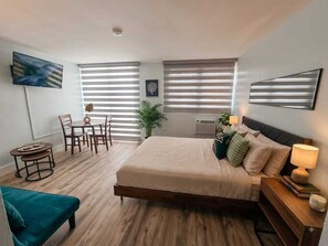 A modern bedroom with a large bed, a desk, and a chair. There are two windows with blinds, a TV mounted on the wall, and a plant on the windowsill. - 🏖Oasis frente al mar en Isla Verde, a minutos del aeropuerto. (Carolina)