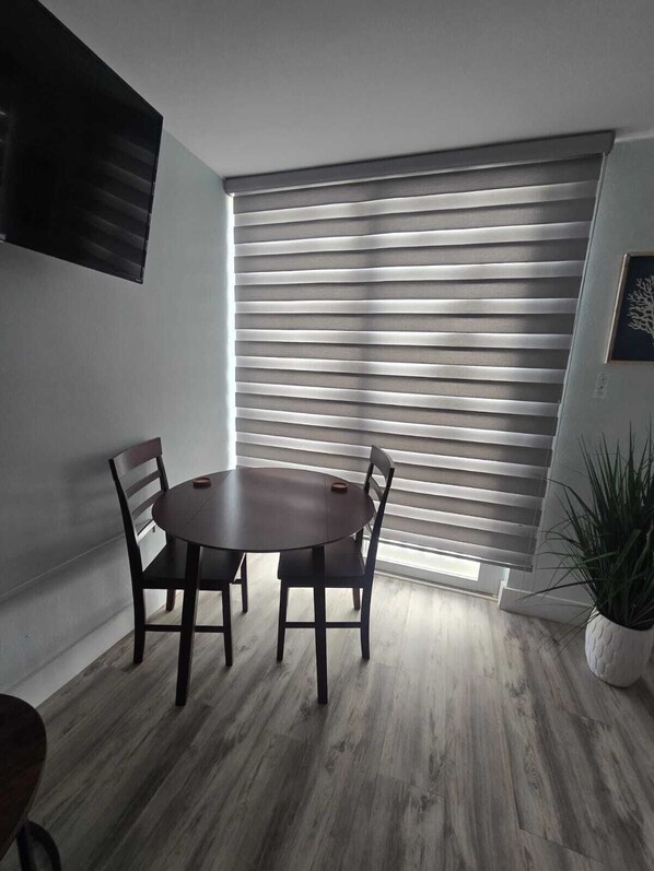 A dining area with a round table and four chairs, a large window with horizontal blinds, a flat-screen TV mounted on the wall, and a potted plant.