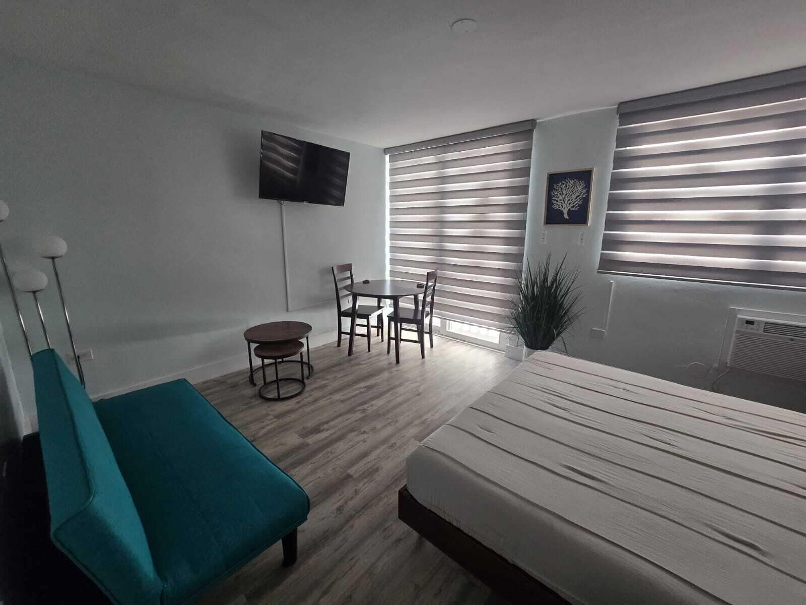 Ein Schlafzimmer mit einem Bett, einer Sitzecke mit Tisch und Stühlen, einem an der Wand befestigten Fernseher und einem Fenster mit Jalousien.