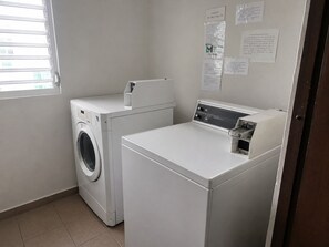 A laundry room with a front-loading washer and dryer, a window with blinds, and informational notices on the wall.