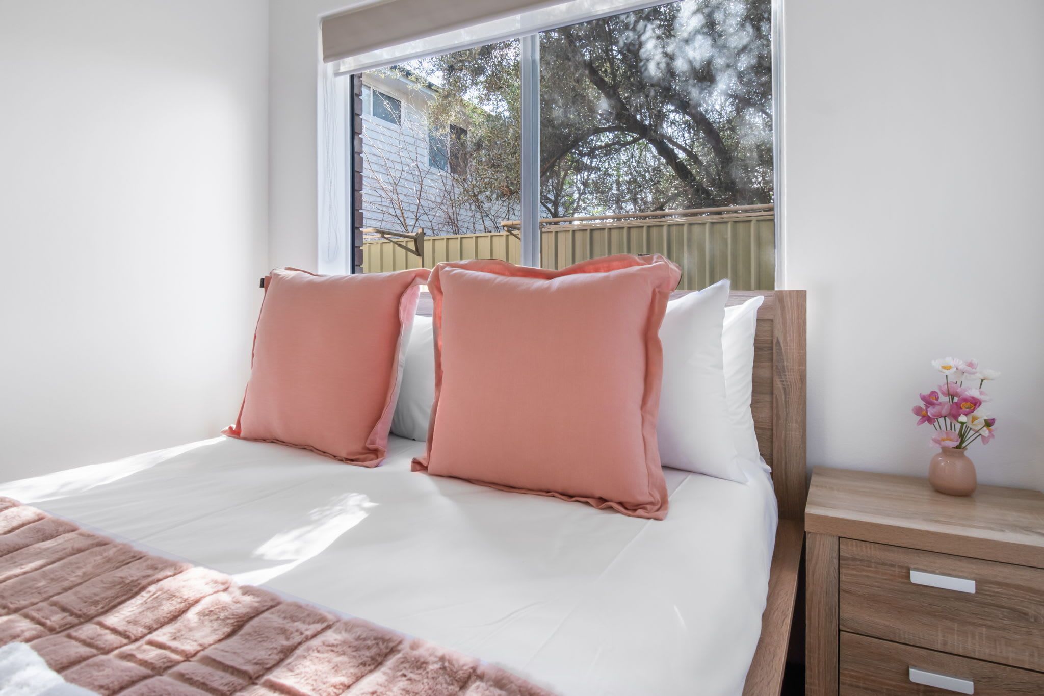 A bedroom with a large window, a bed with pink pillows, a wooden nightstand, and a vase with flowers.