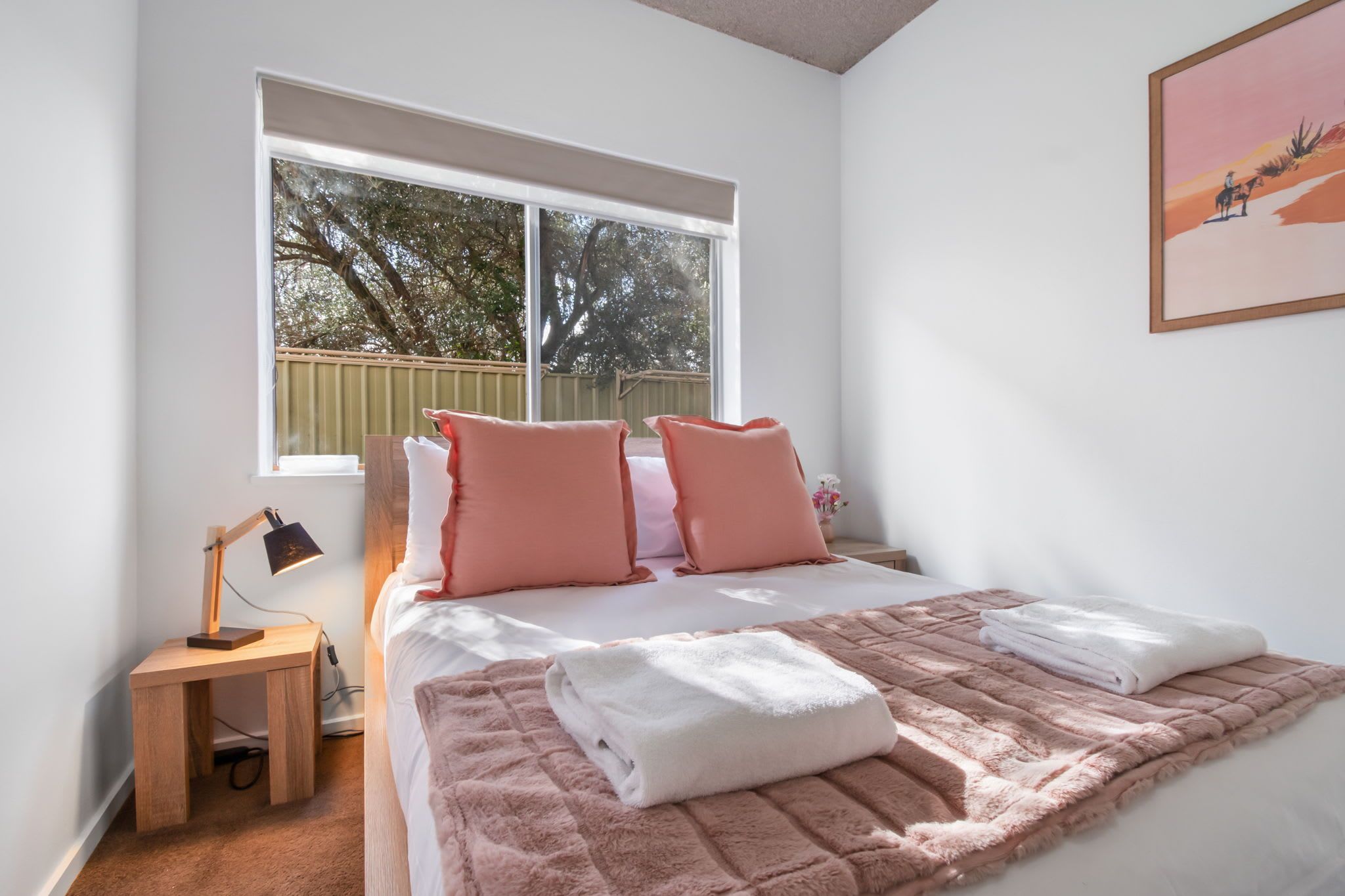 Ein Schlafzimmer mit einem großen Bett, zwei Kissen, einem Nachttisch mit Lampe und einem Fenster mit Blick auf Bäume.
