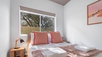 Ein Schlafzimmer mit einem großen Bett, zwei Kissen, einem Nachttisch mit Lampe und einem Fenster mit Blick auf Bäume.