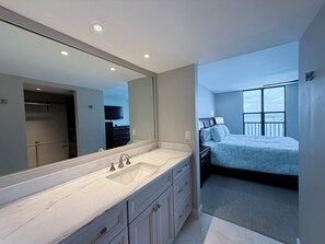 Ein modernes Schlafzimmer mit einem großen Bett, einem Nachttisch und einem Badezimmer mit Waschbecken und Spiegel.