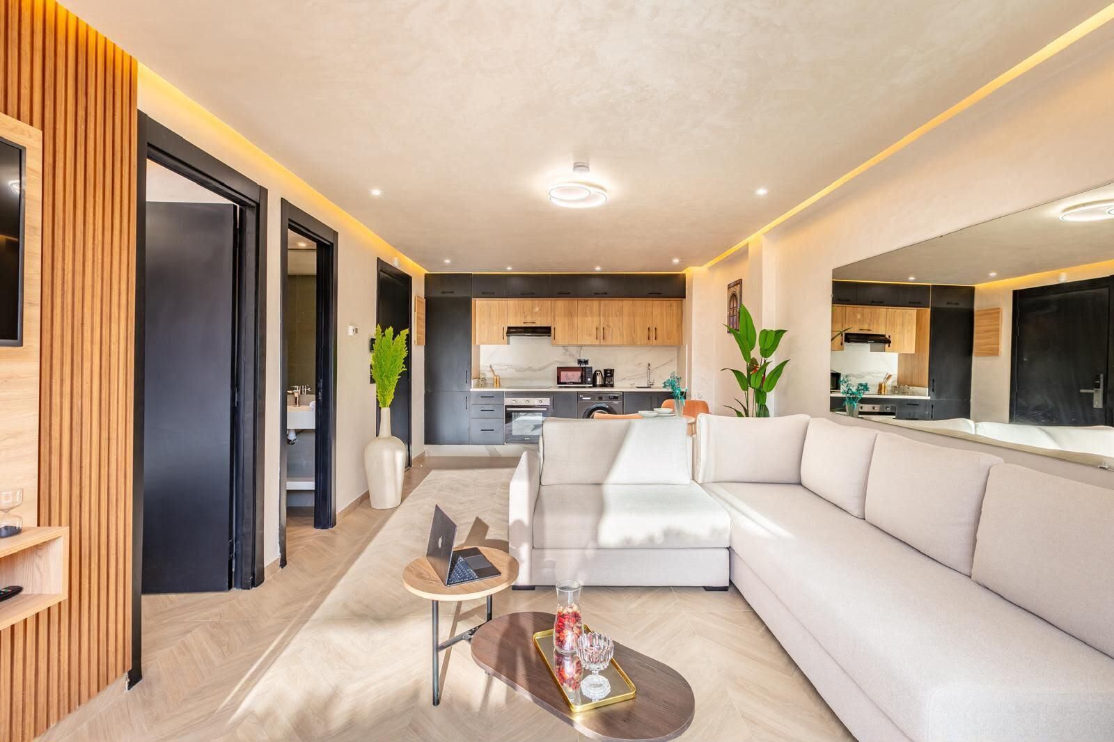 A modern living room with a sofa, a coffee table, and a laptop. There is a kitchen in the background with wooden cabinets and a microwave. - 1-bedroom apartment in tranquil Marrakech with AC (Marrakech)