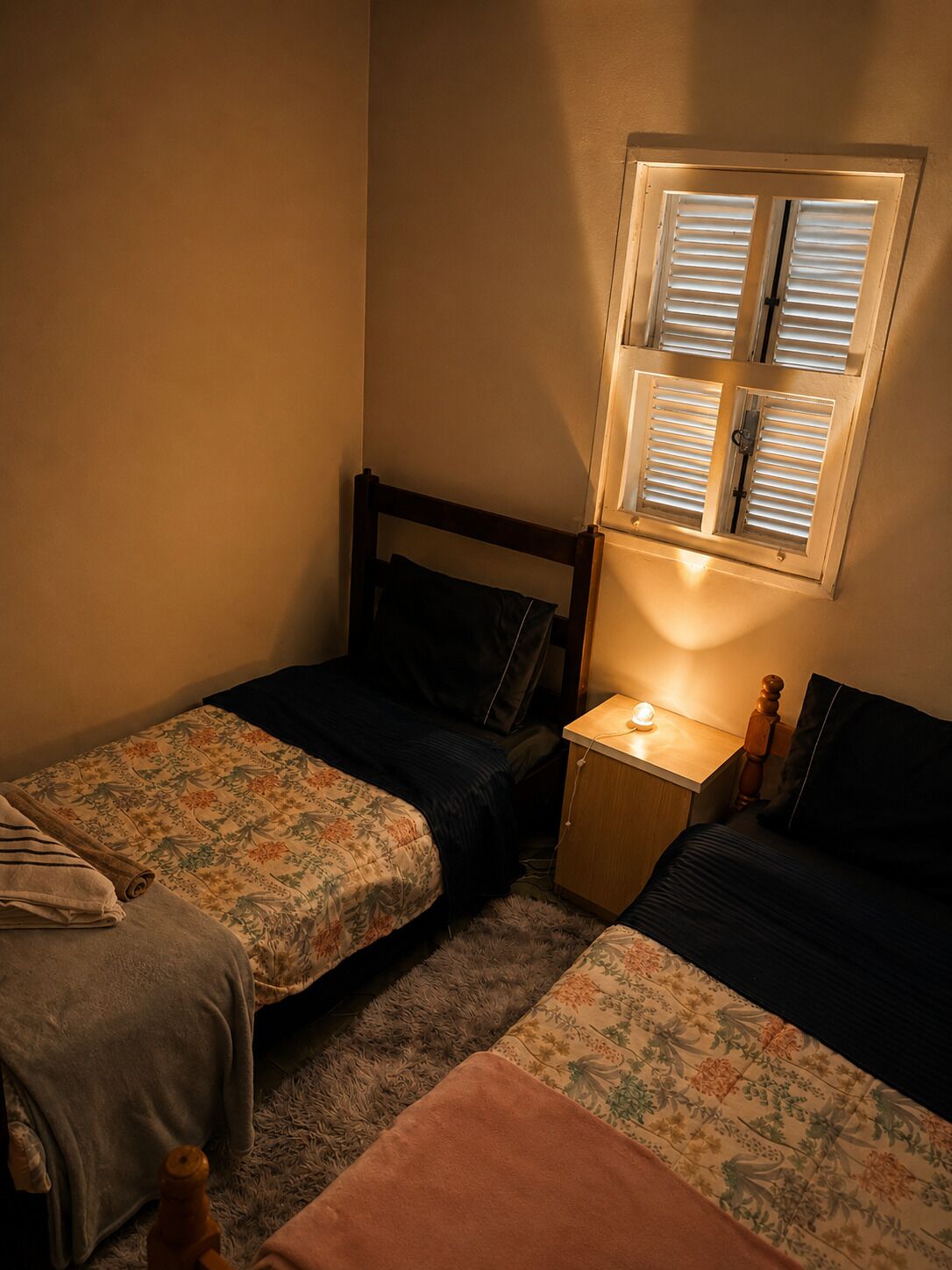 Un dormitorio con dos camas individuales, una mesita de noche con lámpara y una ventana con persianas.