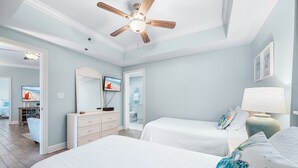 A bedroom with two beds, a ceiling fan, a television, and a bathroom visible through an open door. - Gulf-Front Views | Spacious 3BR | Beach Access & Pool | BEACHBALL PROPERTIES (Orange Beach)