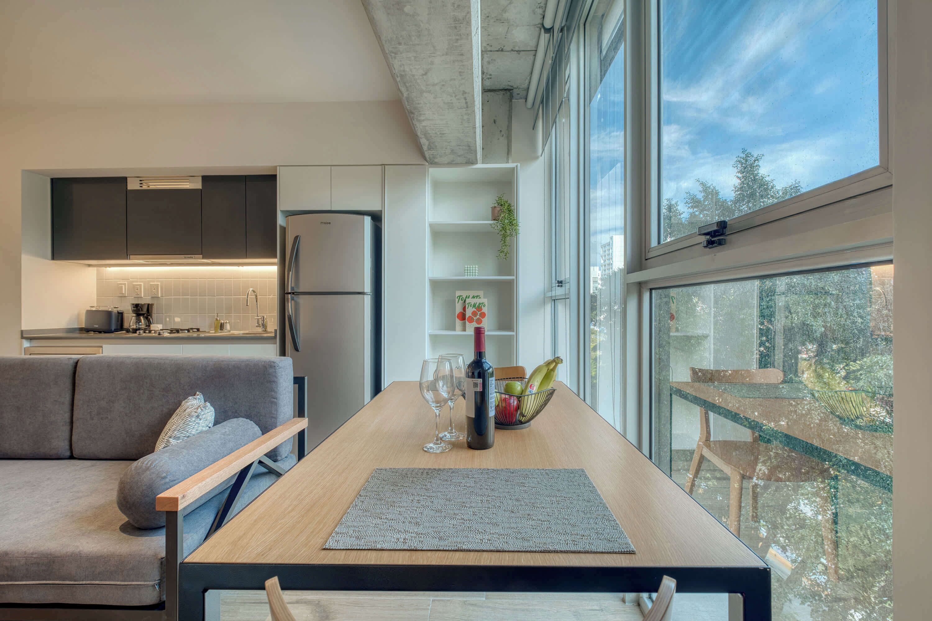 Une cuisine moderne dotée d’un réfrigérateur encastré, d’un coin repas avec une table en bois et d’une vue sur l’extérieur grâce à de grandes fenêtres.