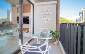 Ein Balkon mit einem Glastisch und Stühlen, einem Blumentopf und einem Regal mit kleinen Pflanzen.