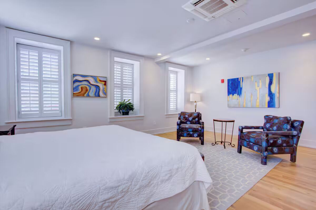 Une chambre d’hôtel avec un grand lit, deux fauteuils, une petite table et des œuvres d’art accrochées aux murs.
