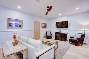 Ein Wohnzimmer mit einem weißen Sofa, einem Holztisch, einem an der Wand befestigten Fernseher und einem Deckenventilator.