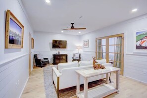 Ein Wohnzimmer mit weißem Sofa, Couchtisch und Deckenventilator. An den Wänden hängen Bilder und es gibt ein Fenster mit Vorhängen.