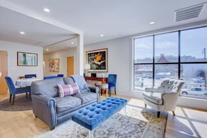 Ein modernes Wohnzimmer mit einem grauen Sofa, einem blauen Hocker und einem weißen Sessel. Es gibt einen Essbereich mit Tisch und Stühlen sowie ein großes Fenster mit Blick auf die Stadt.