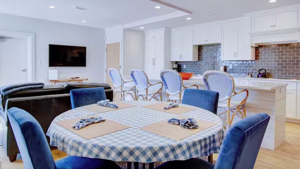 Una cucina moderna con mobili bianchi e una parete a mosaico in mattoni, una zona pranzo con tovaglia a quadri e sedie blu, e un televisore a schermo piatto montato a parete.