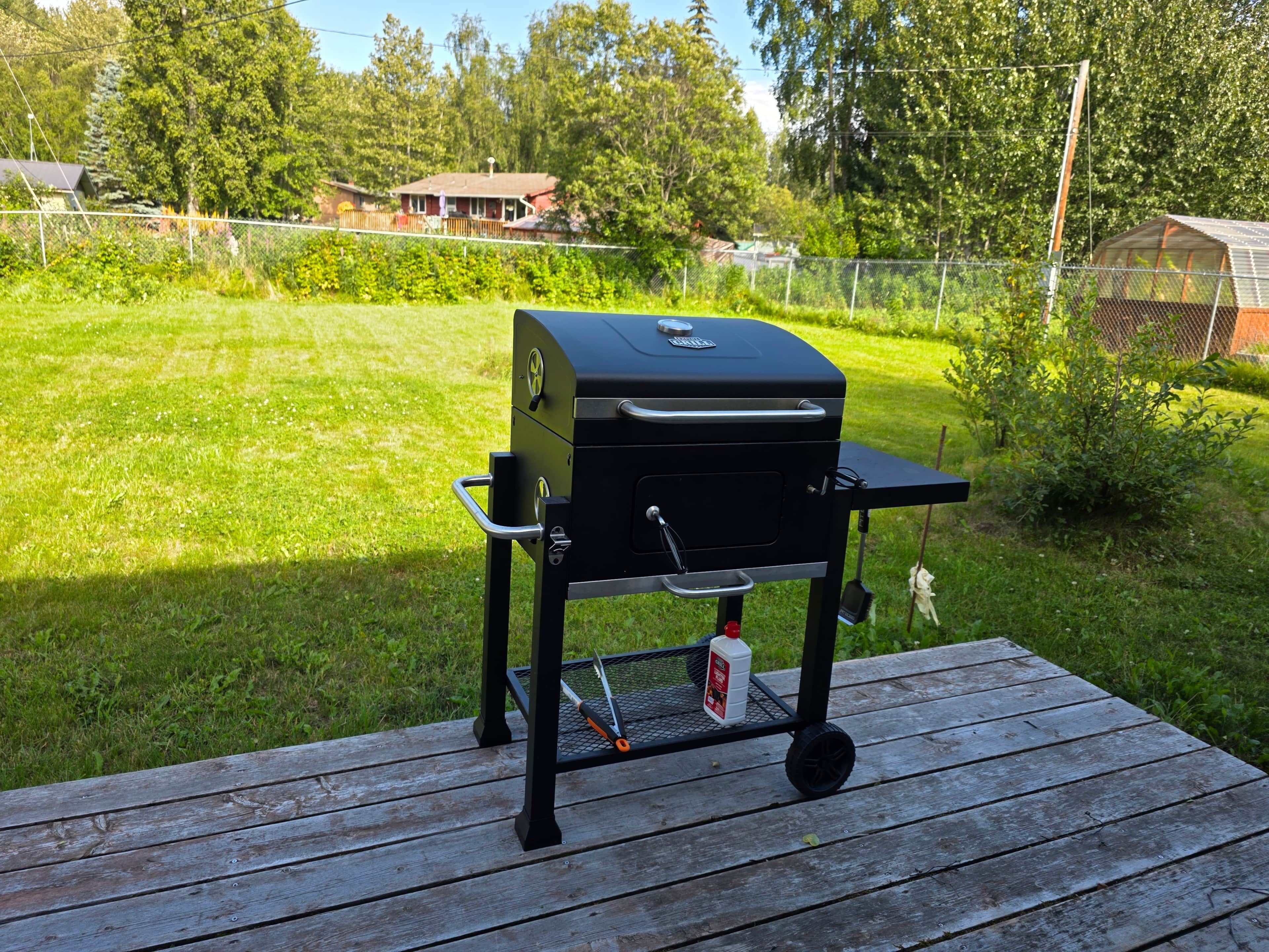 Een draagbare grill op een houten terras, met een gazon en een hek op de achtergrond.