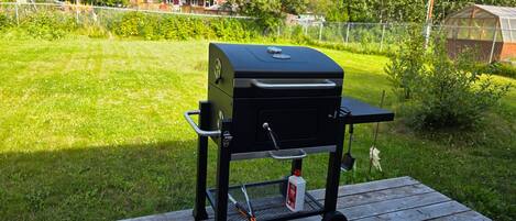 Een draagbare grill op een houten terras, met een gazon en een hek op de achtergrond.