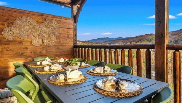 An outdoor dining area with a wooden deck, green chairs, and a table set with plates, bowls, and a centerpiece. - Killer View|Amazing Pool|2 mi Gburg|Game Rm|Modern (Gatlinburg)