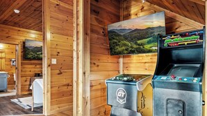 A wooden cabin interior with two arcade machines, a television displaying a landscape, and a small seating area with a table and chairs. - Killer View|Amazing Pool|2 mi Gburg|Game Rm|Modern (Gatlinburg)