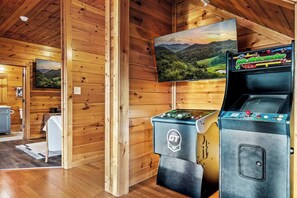 A wooden cabin interior with two arcade machines, a television displaying a landscape, and a small seating area with a table and chairs. - Killer View|Amazing Pool|2 mi Gburg|Game Rm|Modern (Gatlinburg)