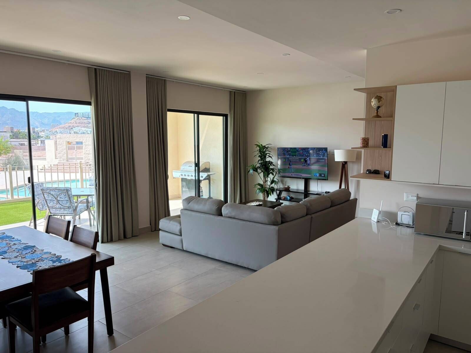 A modern living room with a sofa, a dining table with chairs, a TV, and a view of a pool and mountains.
