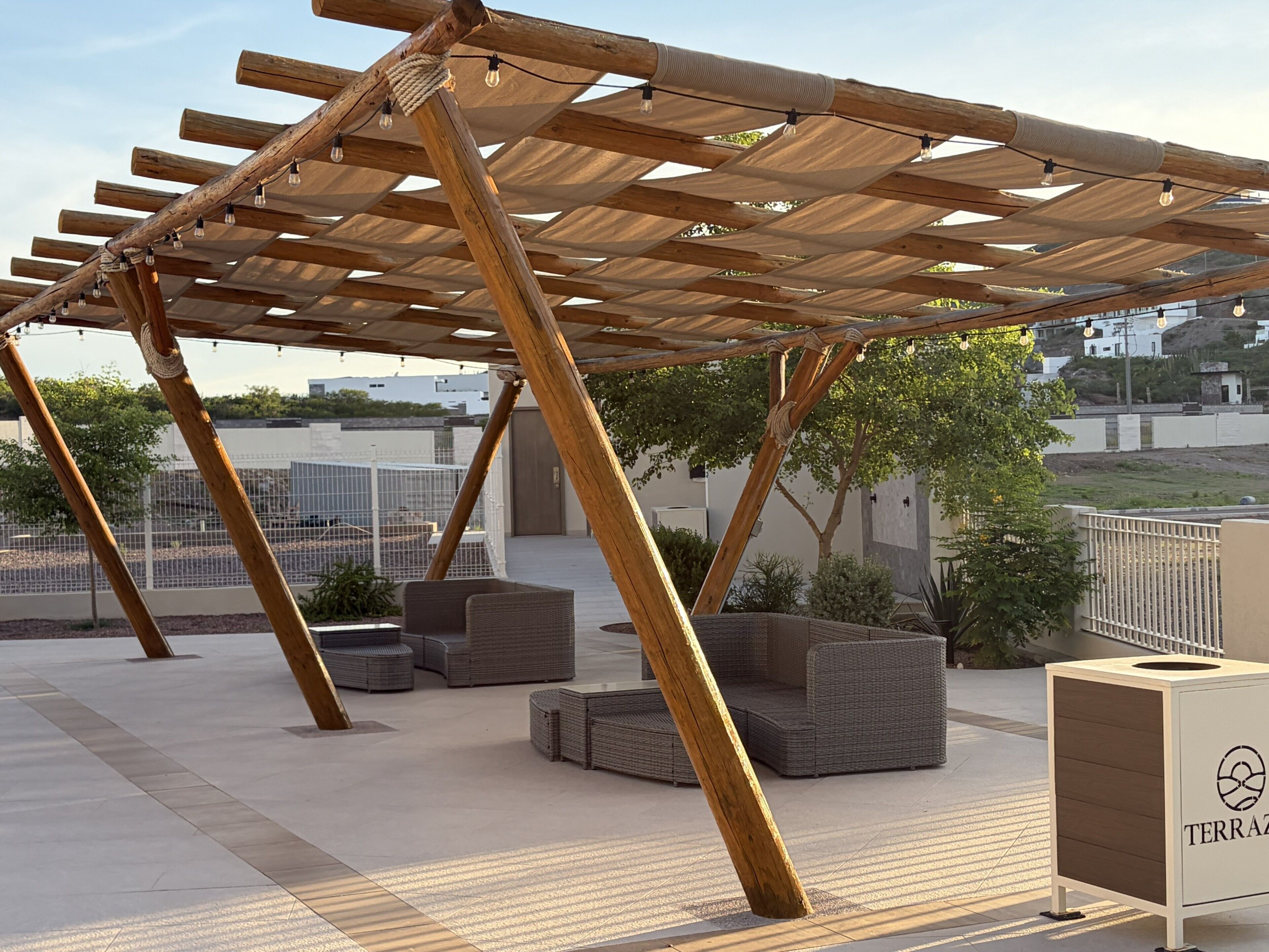 A wooden pergola with light fixtures, outdoor seating, and a branded trash bin.